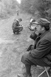ARH NL Mellin 02-102/0012, Jagdhornbläser am Wegrand sitzend, im Hintergrund zwei Männer beim Notieren von Informationen, um 1970