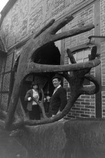 ARH NL Mellin 02-191/0015, Blick durch das Geweih einer Elchskulptur auf ein Ehepaar vor ihrem Bauernhaus, um 1970