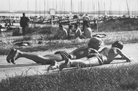 ARH Slg. Bartling 1187, Weisse Düne, 2 Jungen nebeneinander auf dem Radweg liegend und in der Sonne badend, dahinter eine Gruppe weiterer Badegäste und der Bootsanleger, Steinhuder Meer, 1971