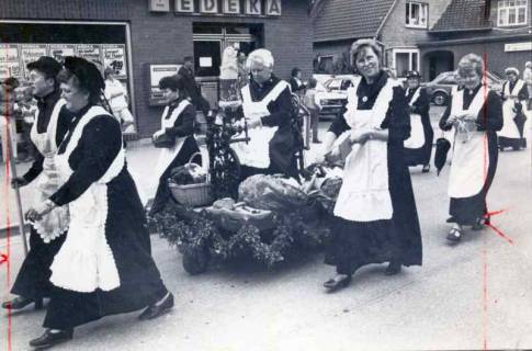 ARH Slg. Bartling 1309, Gruppe von Frauen mit "historischen" Schürzen beim Ernteumzug, in ihrer Mitte eine Karre mit Erntefrüchten, im Hintergrund ein EDEKA-Laden, Mandelsloh, 1984