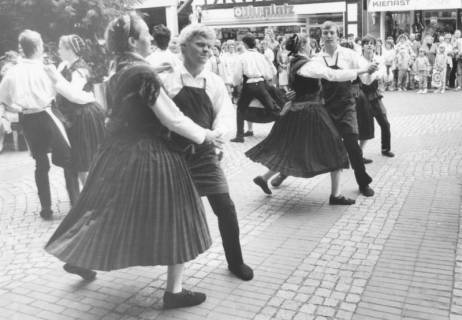 ARH Slg. Bartling 1770, Volkstanzgruppe aus sechs Paaren in Tracht (Damen mit Faltenrock, gehäkeltem Schultertuch und Kappe, Herren mit weißem Hemd und Handwerkerschürze) bei einer Vorführung auf dem Platz vor der Kreissparkasse, Neustadt a. Rbge., um 1975