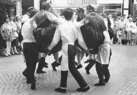 ARH Slg. Bartling 1782, Volkstanzgruppe aus sechs Paaren in Tracht (Damen mit Faltenrock, gehäkeltem Schultertuch und Kappe, Herren mit weißem Hemd und Handwerkerschürze) bei einer Vorführung auf dem Platz vor der Kreissparkasse, Neustadt a. Rbge., um 1975