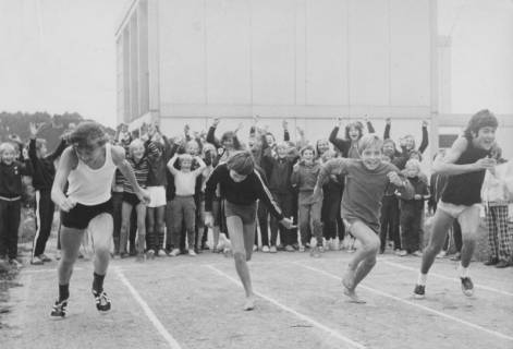 ARH Slg. Bartling 1947, Vier Jungen beim Start zum Wettlauf auf einem Sportplatz, im Hintergrund eine Gruppe von anfeuernden Schülern vor einem zweigeschossigen Gebäude aus Betonfertigteilen; Blick von vorn, Neustadt a. Rbge., 1971