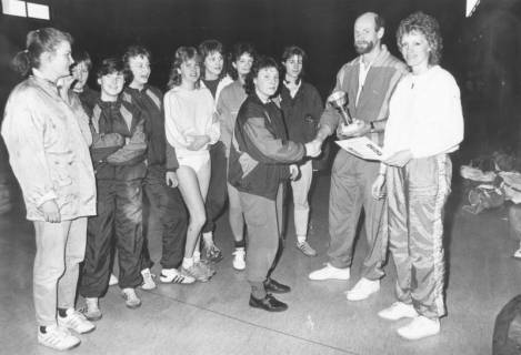 ARH Slg. Bartling 2038, Überreichung eines Pokals und einer Urkunde an junge Hallenhandballerinnen (?) durch einen Mann und eine Frau in einer Turnhalle, um 1975