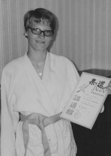 ARH Slg. Bartling 2073, Einzelporträt einer jungen Frau (S. Wiskemann) als Karateka im Karate-Gi mit hellem Gürtel, Brille tragend, eine Urkunde mit der Linken zeigend, in häuslicher Umgebung, um 1975