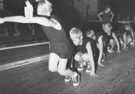 ARH Slg. Bartling 2125, Gruppe von Kindern bei einer Grundübung in einer Turnhalle hintereinander in Hockstellung hüpfend, Neustadt a. Rbge., 1972