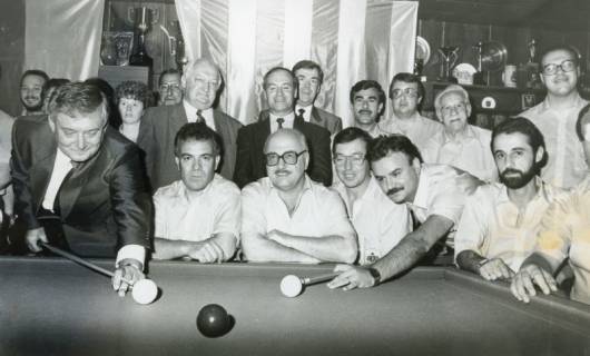ARH Slg. Bartling 2249, Zwei Billardspieler (l. Günther Paschke, r. N. N.), umgeben von zahlreichen sitzenden und stehenden Zuschauern (darunter Bürgermeister Henry Hahn [linke Mitte]), beim Spiel am Tisch im Billardstudio, Neustadt a. Rbge., um 1985