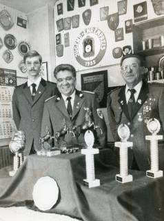 ARH Slg. Bartling 2310, Drei uniformierte Sportschützen hinter einem Tisch mit unterschiedlichen Trophäen (darunter springende Pferde) vor einer Wand mit vielen Plaketten und Ehrenzeichen (darunter ein "Jugendehrenpreis 1985"), Neustadt a. Rbge., um 1985