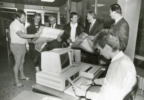 ARH Slg. Bartling 2418, Im Hallenbad eine junge Frau am PC sitzend und schreibend, dahinter im Halbkreis stehend eine Gruppe von sechs Männern die ein Werbeblatt der Barmer Ersatzkasse studieren, Neustadt a. Rbge., zwischen 1970/1972