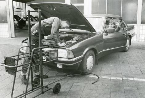 ARH Slg. Bartling 2581, Überprüfung der Scheinwerfer eines Fahrzeugs (mit geöffneter Motorhaube) durch zwei junge Männer vor dem Werkstatt Gebäude vom Autohaus A. W. Lange Ford-Händler, Nienburger Straße 1, Neustadt a. Rbge., zwischen 1981/1985