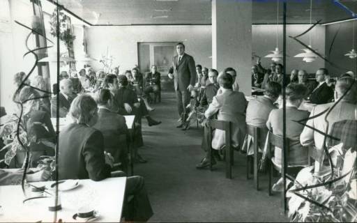 ARH Slg. Bartling 3162, Kreiskrankenhaus (später: KRH Klinikum Neustadt a. Rbge.), Lindenstraße 75: Referat des Verwaltungsdirektors Friedrich Schmidt (später Manhal) (stehend) vor an Tischen sitzenden Männern und Frauen im Aufenthaltsraum, Neustadt a. Rbge., 1974