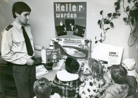 ARH Slg. Bartling 3370, Belehrung von Kindergartenkindern durch einen jungen Polizisten (l.), Blick über die Hinterköpfe der Kinder auf einen Tisch mit Lernmaterial, Neustadt a. Rbge., nach 1976