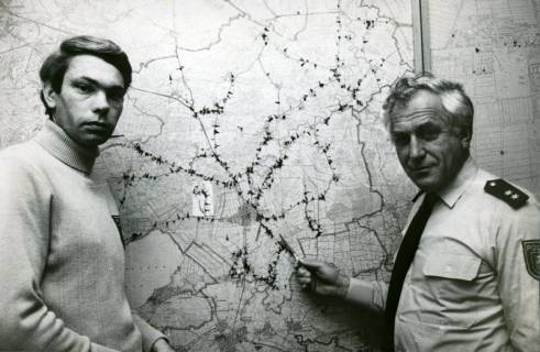 ARH Slg. Bartling 3381, Polizeihauptkommissar Strasser (r.) und Polizist Overhey (?) beim Studium einer an der Wand hängenden topografischen Karte von Neustadt mit Eintragung der Unfallschwerpunkte, Neustadt a. Rbge., um 1975