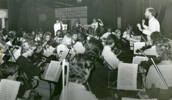 ARH Slg. Bartling 3671, Aufführung des Schulorchesters des Gymnasiums (?) unter der Leitung von N. N., Blick von der linken Seite, im Hintergrund auf der Bühne die Kulisse eines Hauses, Neustadt a. Rbge., um 1985