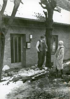 ARH Slg. Bartling 3690, Mann und Frau vor einem Haus mit der Nr. 29, an dessen Haustür das winterliche Hochwasser heranreicht, um 1985