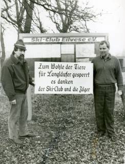 ARH Slg. Bartling 3693, Zwei Männer (Godau (li.) und Duensing) vor dem Schaukasten des Ski-Clubs Eilvese e. V. stehend, zwischen sich ein Schild haltend mit der Aufschrift "Zum Wohle der Tiere für Langläufer gesperrt ...", Eilvese, um 1985