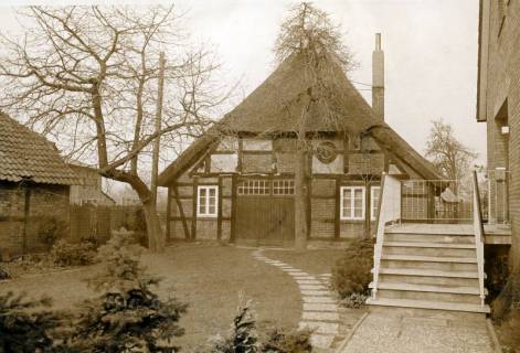 ARH Slg. Bartling 4674, Bauernhaus, Fachwerkbau mit Krüppelwalmdach, Strohdach, Giebelansicht mit Rasen-bedecktem Vorhof, rechts die Zugangstreppe zu einem modernen Wohnhaus, Zuwegung über Waschbetonplatten, Niedernstöcken, um 1975