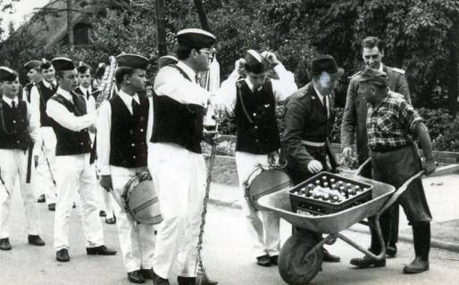 ARH Slg. Bartling 4687, Stopp des Spielmannszugs (unter der Leitung von Gerhard Knuth?) beim Marsch auf der Straße durch einen Mann mit einem Kasten Bier auf einer Schubkarre beim Schützenfest, Niedernstöcken, 1969