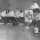 ARH Slg. Bartling 2089, Versammlung von jungen Turner*innen in einer Turnhalle auf Bänken im Halbkreis sitzend und der Rede des Geschäftsführers des Deutschen Turnerbundes Erich Heinz Ertl (an einem mit dem Turnerkreuz geschmückten Pult) zuhörend