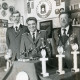 Stadtarchiv Neustadt a. Rbge., ARH Slg. Bartling 2310, Drei uniformierte Sportschützen hinter einem Tisch mit unterschiedlichen Trophäen (darunter springende Pferde) vor einer Wand mit vielen Plaketten und Ehrenzeichen (darunter ein "Jugendehrenpreis 1985"), Neustadt a. Rbge.