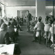 Stadtarchiv Neustadt a. Rbge., ARH Slg. Bartling 3162, Kreiskrankenhaus (später: KRH Klinikum Neustadt a. Rbge.), Lindenstraße 75: Referat des Verwaltungsdirektors Friedrich Schmidt (später Manhal) (stehend) vor an Tischen sitzenden Männern und Frauen im Aufenthaltsraum, Neustadt a. Rbge.