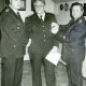 Stadtarchiv Neustadt a. Rbge., ARH Slg. Bartling 3681, Ehrung von Eduard Formella durch Überreichung einer Urkunde und eines Anhängers in einem Dachzimmer durch Stadtbrandmeister Horst Liem (r.) und Ortsbrandmeister Heinrich Lammers (l.) im Gerätehaus, An der Beeke, Mariensee
