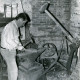 Stadtarchiv Neustadt a. Rbge., ARH Slg. Bartling 4263, Vorsitzender des Schmiedevereins Helstorf N. N. in Freizeitkleidung beim Versuch, mit dem Hammer am Amboss ein Stück Eisen zu formen in der Alten Schmiede, Helstorf