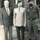 Stadtarchiv Neustadt a. Rbge., ARH Slg. Bartling 4334, Gruppenporträt der drei Männer Herbert Schöling (in Zivil), Schmelzer (in Dienstuniform) und Kompaniechef Busch (in Arbeitsuniform, mit Vollbart) vor einem Kasernengebäude mit Wappenschild, Luttmersen