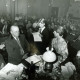 ARH Slg. Bartling 4428, Gut besuchte Versammlung im Saal des Gasthauses Küster, Blick aus der Mitte des Saales auf den Tisch von Honoratioren, darunter eine jüngere Lehrerin (?), neben der ein Junge mit Mikrofon einen Text vom Blatt abliest, Mandelsloh