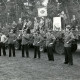 Stadtarchiv Neustadt a. Rbge., ARH Slg. Bartling 4554, Auftritt einer Gruppe von Jagdhornbläsern auf einer Wiese am Waldrand vor einem Wagen mit Festtagsrednern und VIPs beim Kreis-Schützenfest (2 Ex.), Mardorf