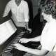 Stadtarchiv Neustadt a. Rbge., ARH Slg. Bartling 4686, Junge Frau sitzend beim Spiel auf einer elektronischen Orgel,  am Ende stehend und zuhörend ein junger Mann, Niedernstöcken?