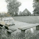 Stadtarchiv Neustadt a. Rbge., ARH Slg. Bartling 4738, Gedenkstein (Findling) am Friedhof Nöpke 1873-1955, Blick über das eingeebnete Areal auf drei übrig gebliebene Steinsäulen und den Giebel eines am Rande stehenden Hauses (2 Ex.), Nöpke