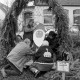 Archiv der Region Hannover, ARH Slg. Weber 02-002/0009, Personen stellen eine Baumstamm mit einem Weihnachtsmann-Gesicht unter einem Tannenbogen auf, im Hintergrund das Dorfgemeinschaftshaus, Redderse